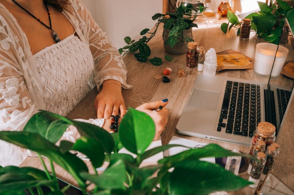 A peaceful workspace featuring a woman writing, surrounded by greenery, crystals, and a laptop.